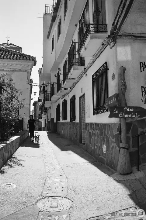 A black-and-white photo of a narrow street in Soportújar with a lady walking her dog. St. María la Mayor church is on the left