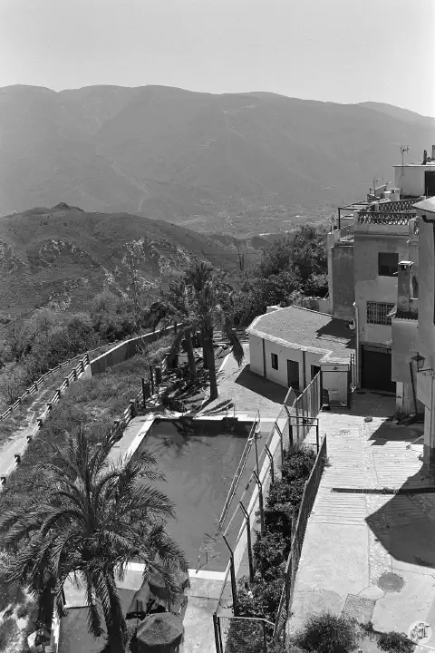 A black-and-white photo of the view from Mirador del Embrujo. Below is a swimming pool and white houses, with hills and mountains in the distance.