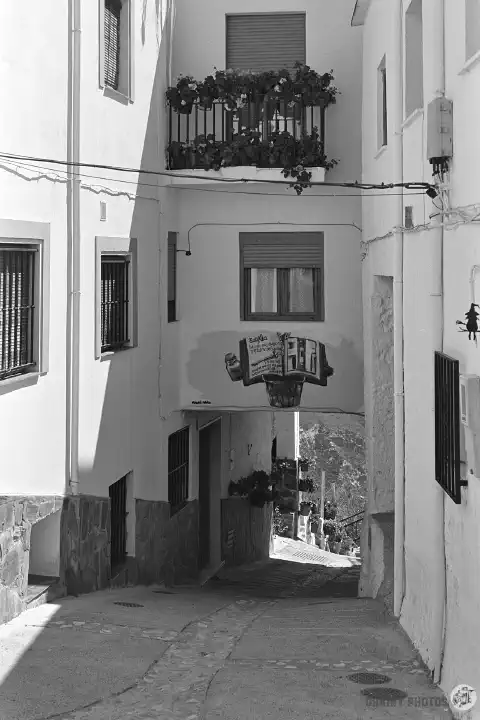 A black-and-white photo of a narrow street in Soportújar with a mural of a witch's spell book on the wall.