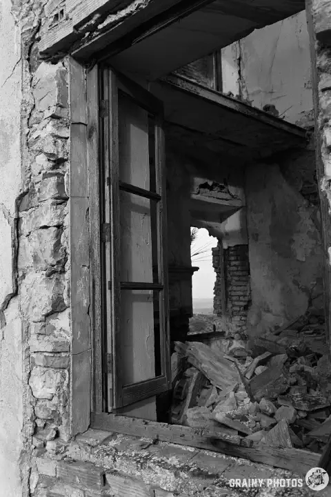 A black-and-white photo looking through another window. Part of the window frame is still in place. The first floor has collapsed, and there is a lot of rubble inside.