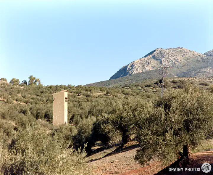 A colour film photo of a rendered tower-like structure with power lines running to an electrical power pylon. This is amongst the olive groves with a mountain in the distance.
