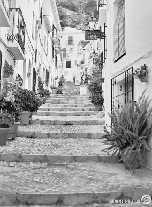 A narrow, winding stone staircase leads up through whitewashed buildings adorned with potted plants. The scene captures a charming, serene atmosphere, evoking a sense of exploration and nostalgia.