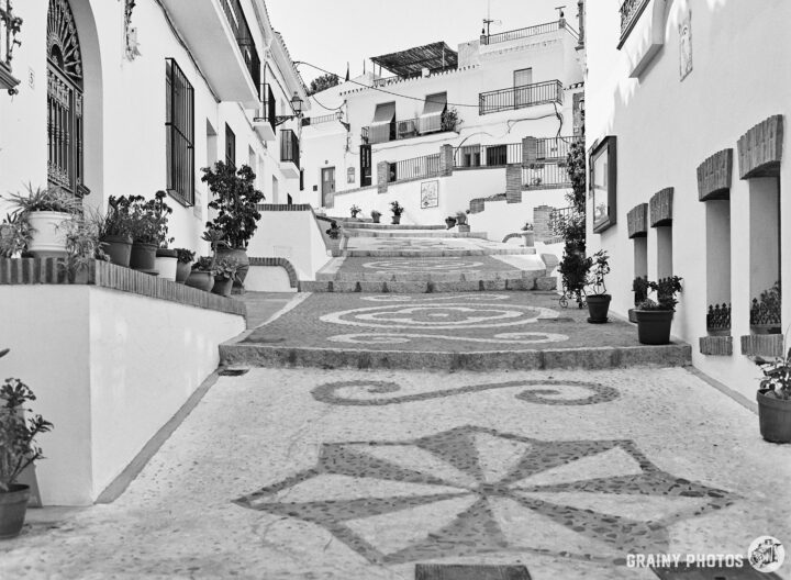 A charming cobblestone street with flower pots lining the path, leading up to white-washed houses in a picturesque hillside village. The scene is enhanced by intricate patterns in the pavement, creating a serene and inviting atmosphere.