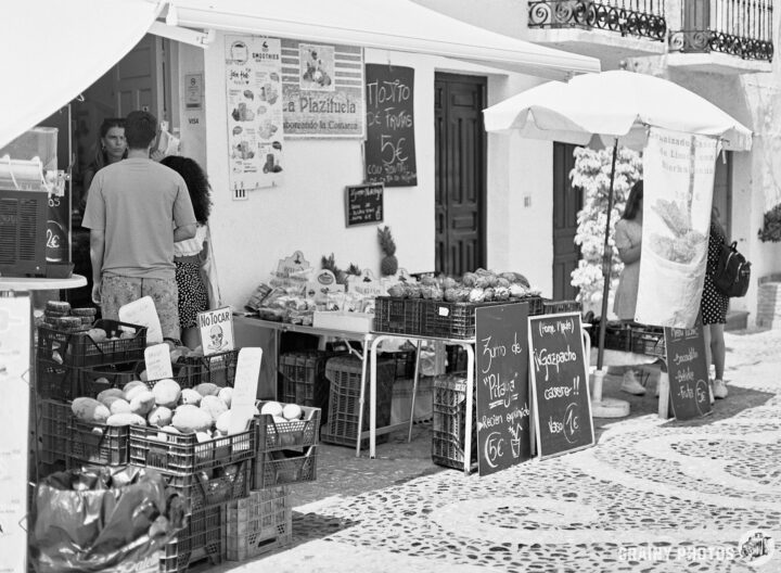 A black and white photo of a small village shop selling fresh produce, including fruits and vegetables. Customers interact at the entrance, with signs displaying prices for various items. The setting is picturesque.