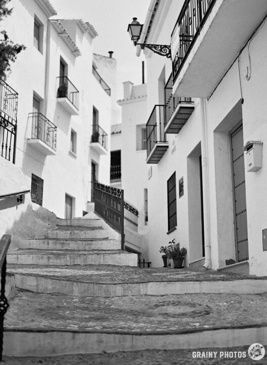 A narrow, winding stepped alley leads up through Frigiliana, flanked by charming whitewashed buildings with balconies, set against a clear sky.
