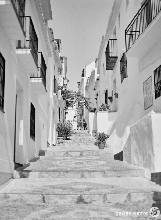 A narrow, winding street ascends through whitewashed buildings adorned with balconies, leading to a vibrant scene with potted plants and a couple walking together, capturing the charm of a quaint village in black and white.