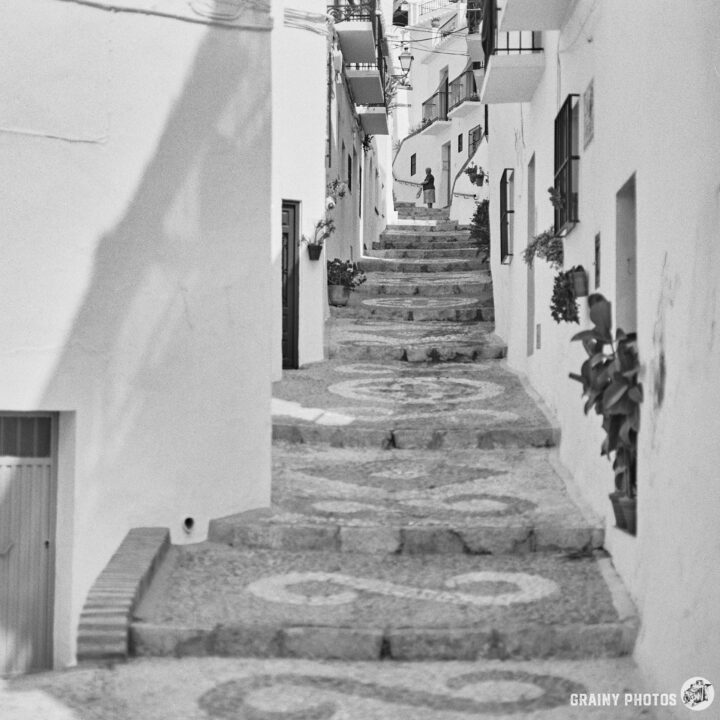 A narrow cobblestone alleyway lined with white buildings, featuring decorative planters. A person walks up the winding steps, surrounded by a charming, picturesque atmosphere.