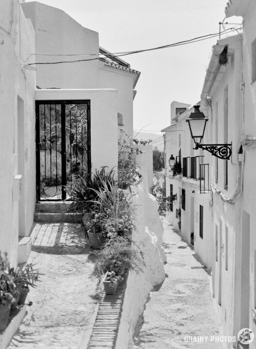 A narrow, winding alleyway lined with whitewashed buildings. A decorative lantern hangs above a gate, with potted plants adding greenery. The scene conveys a tranquil, historic atmosphere in a charming urban setting.