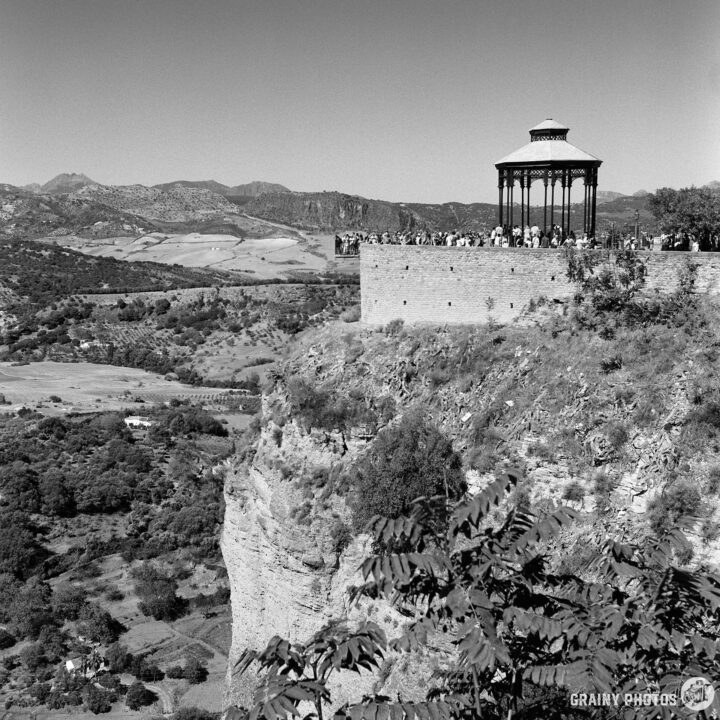 A scenic view of a cliffside Mirador in Ronda overlooking a lush valley, with people enjoying the landscape. The image features rolling hills and distant mountains, captured in black and white.