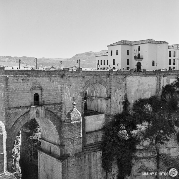 The historic Puente Nuevo in Ronda, Spain, with stone arches, surrounded by lush greenery and residential buildings. The scene captures the architectural beauty against a clear sky, showcasing the stunning landscape and cultural heritage of the area.