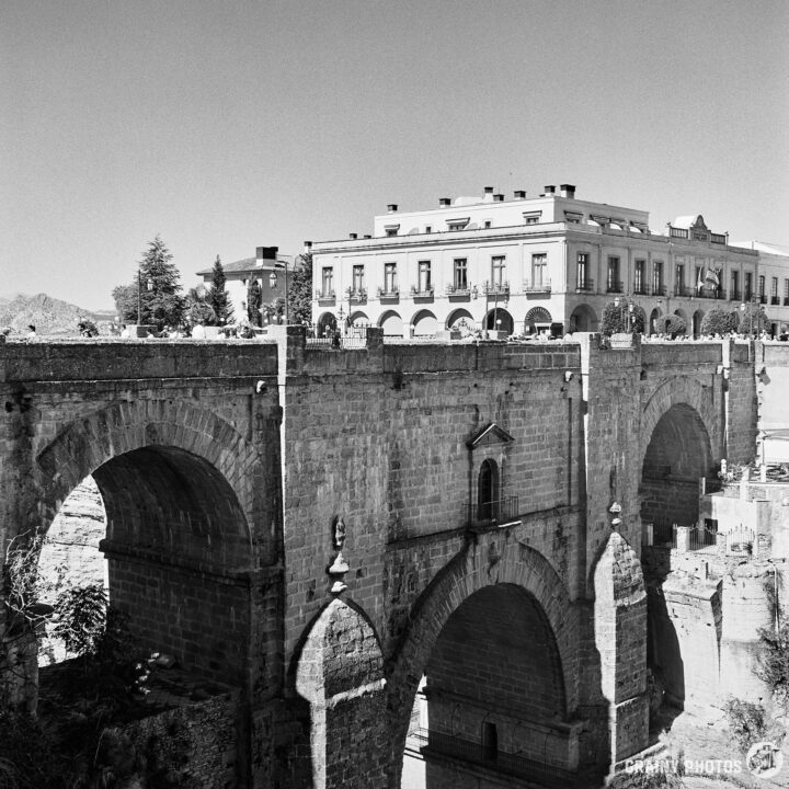 Black and white image of the Puente Nuevo. A grand building with arches stands on the far side. The scene captures architectural elegance and the charm of Ronda.