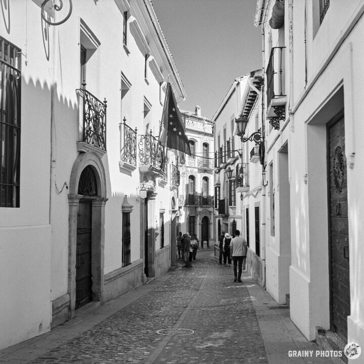A narrow street in the old town, featuring traditional architecture and ornate balconies. A European flag hangs from a building as people stroll along the cobblestone path, enjoying the sunny day.