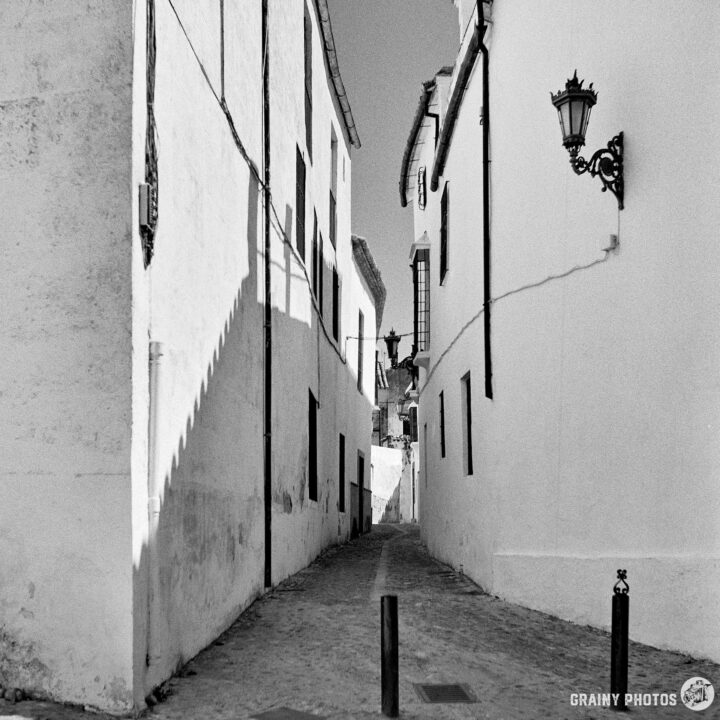 A narrow, cobblestone alleyway between white-washed buildings with shadows cast along the walls, featuring vintage-style street lamps, evoking a timeless charm and quiet ambiance.