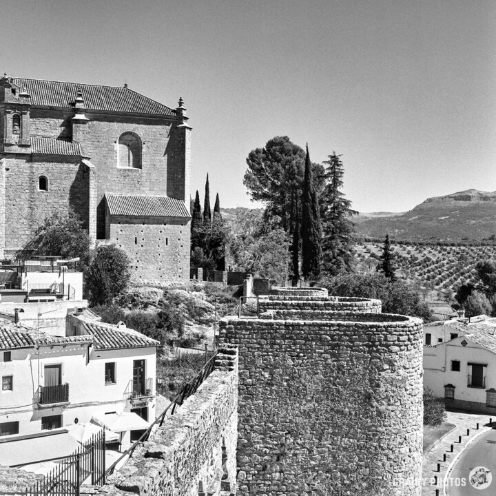 Restored city walls and a prominent church, surrounded by trees and hills, set against a clear sky. The image captures architectural details and the surrounding landscape in black and white.