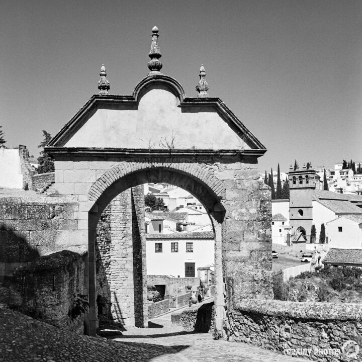 Arco de Felipe V leading to white buildings and a visible church tower in the background, framed by trees and a clear sky.