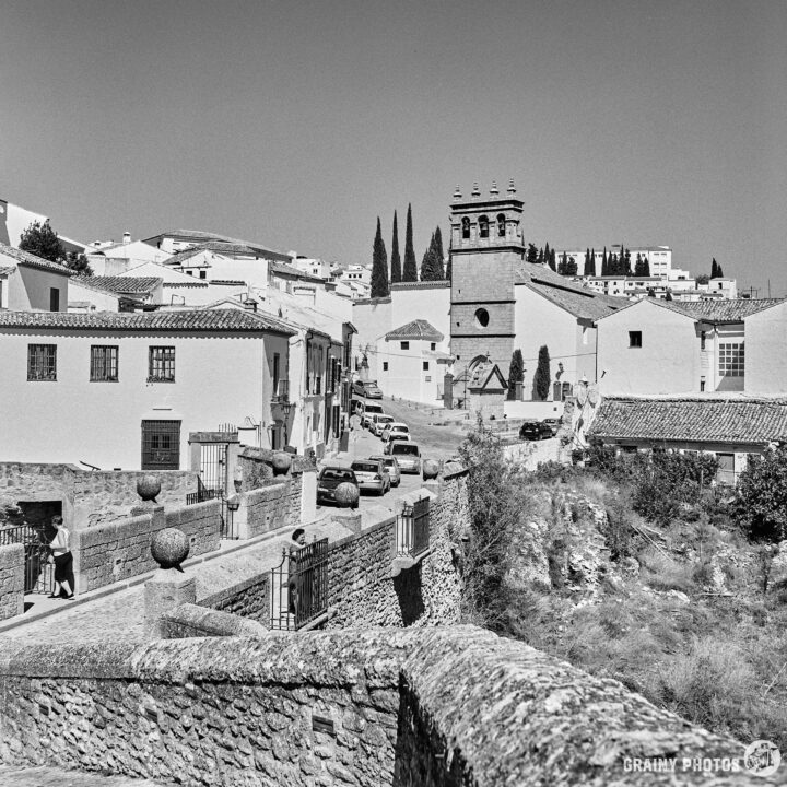 A serene black-and-white image of the newer part of the city with narrow streets, whitewashed buildings, and a prominent clock tower. Lush greenery and cypress trees are visible in the background, enhancing the peaceful atmosphere of this picturesque Spanish town.