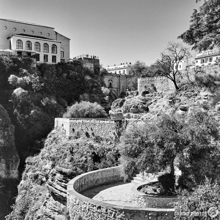 Footpath and viewing terraces on the side of el Tajo Gorge featuring steep cliffs, terraced gardens, and a circular stone pathway, with a building visible in the background and sparse trees dotting the terrain.