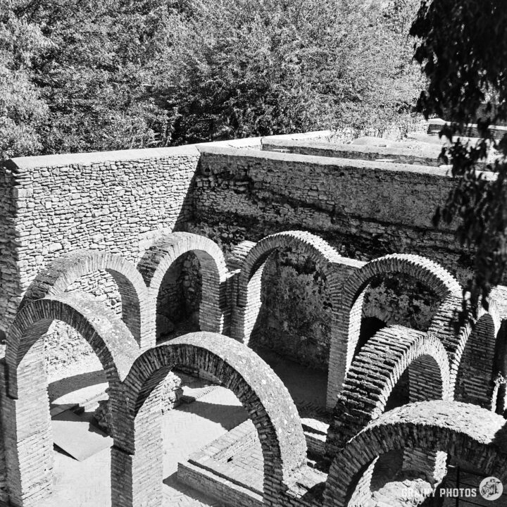 Black and white image of Baños Árabes ruins featuring arched structures and a lush tree backdrop. The arched designs emphasize the architectural elements, creating a striking contrast with the surrounding nature.