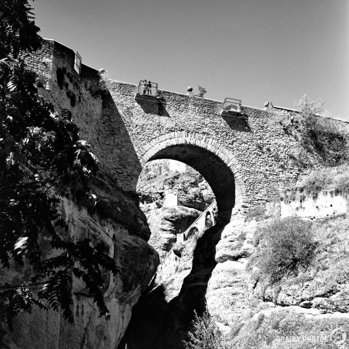 A black and white photograph of Puente Viejo, a stone arch bridge spanning a ravine, surrounded by rocky terrain and vegetation. The bridge is elevated, with stairs leading down to the ravine, conveying a sense of historical architecture and natural beauty.