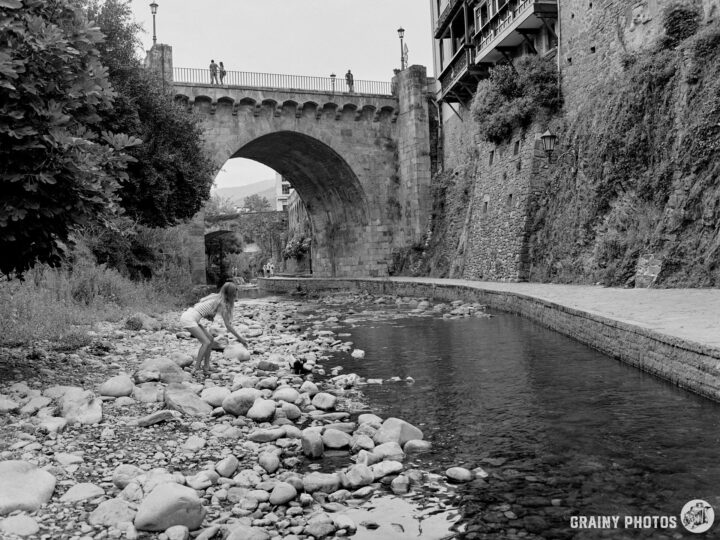 A child feeds ducks in a riverbed filled with stones, near an old stone bridge. Lush greenery surrounds the area, with people walking on the bridge above and along the riverbank.