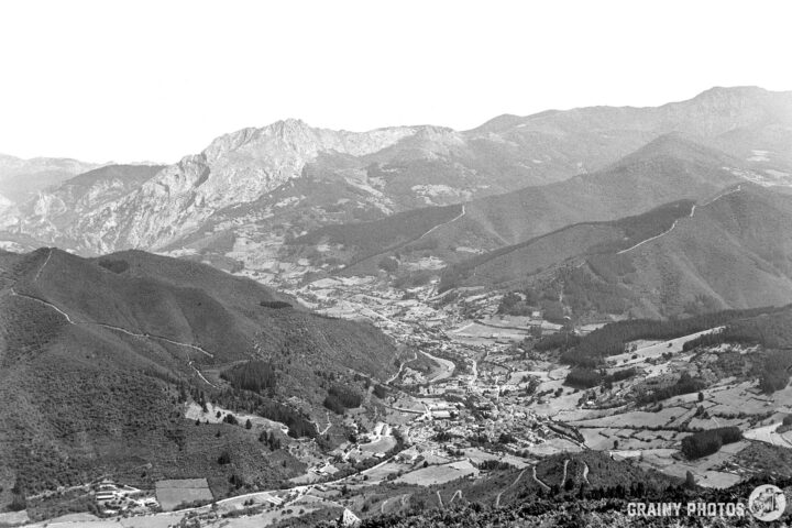 A black and white panoramic view of Potes nestling in the Liébana valley surrounded by a mountainous landscape. Winding roads cut through the terrain, creating a serene and picturesque scene.