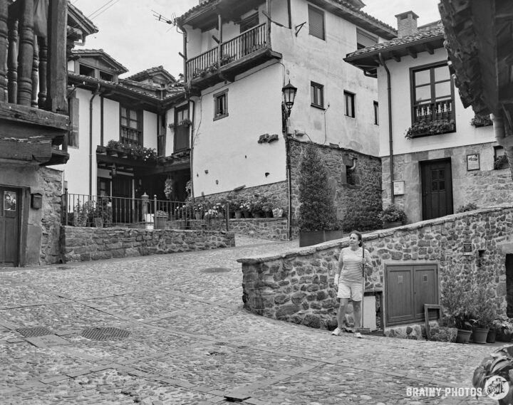 A woman with a broom stands in a quaint, narrow cobblestone street surrounded by rustic buildings adorned with balconies and greenery, embodying the charm of a traditional village setting in black and white.