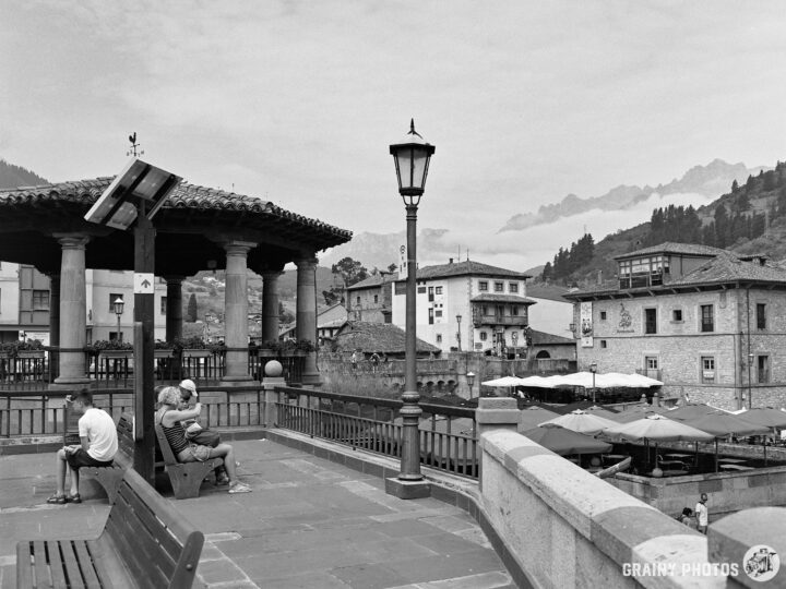A black and white view of a plaza with benches, a lamp post, and nearby buildings. People are seated, enjoying the scenery, while misty mountains are visible in the background, creating a serene atmosphere.
