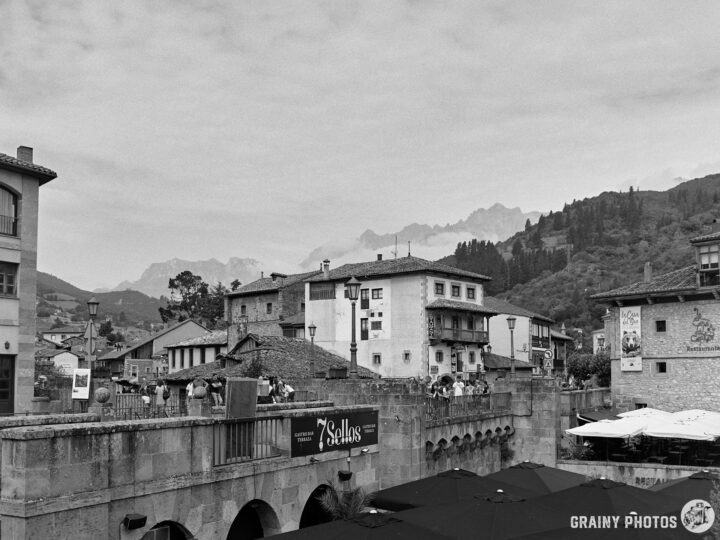 A scenic view of Potes with historical buildings, nestled between mountains under a cloudy sky. People gather on a picturesque bridge, with tables and umbrellas visible, creating a lively atmosphere in this black-and-white photograph.