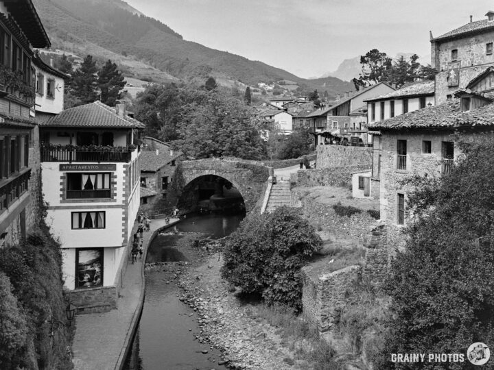 A scenic view of Potes, featuring old stone buildings, a quaint arch bridge, and a gently flowing river, surrounded by lush greenery and mountains in the background, captured in a black and white tone.
