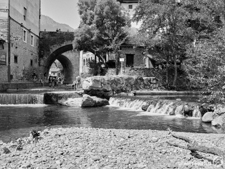 A peaceful black-and-white scene featuring a gentle waterfall flowing into a rocky riverbed, with a stone arch bridge in the background surrounded by trees and quaint buildings. People can be seen enjoying the serene environment.