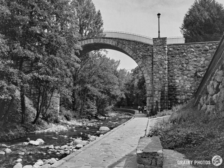 A black-and-white image of a stone bridge arching over a river, surrounded by trees. A pathway runs alongside the water, with smooth stones lining the shore, creating a serene natural landscape.