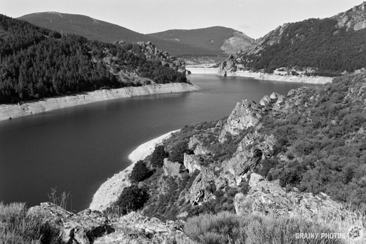 A scenic black and white view of a reservoir meandering through rocky terrain surrounded by lush forests and distant mountains. The landscape captures the tranquility of nature, highlighting the contrasts between water, rocks, and greenery.