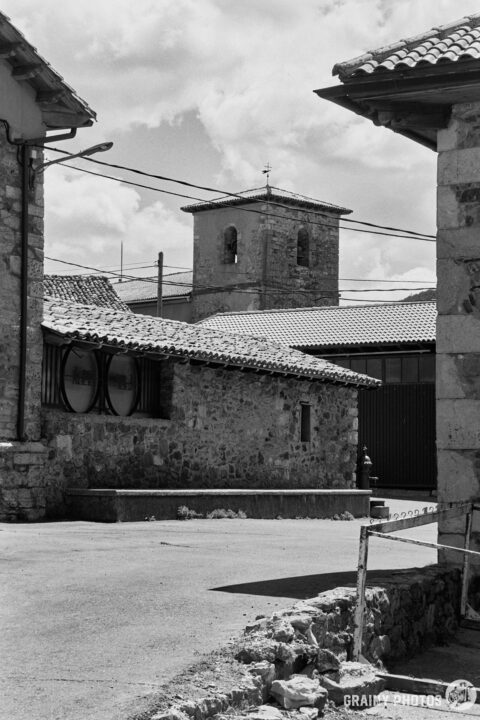 A black and white photograph depicting a picturesque village scene, featuring stone buildings, a bell tower in the background, and a winding street under a cloudy sky.
