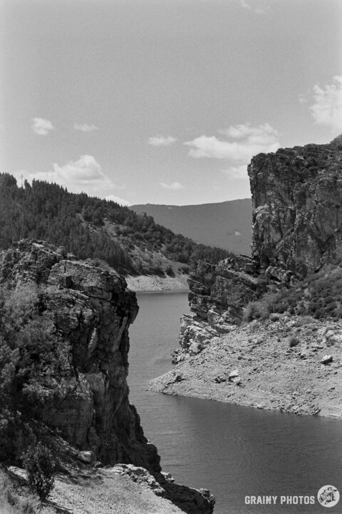 A scenic view of the Camporredondo Reservoir between rocky cliffs under a mostly cloudy sky, surrounded by dense greenery. The landscape captures the natural beauty of the area in a timeless black and white format.