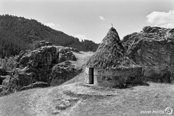 A small, thatched shepherd’s hut sits near rocky outcrops, surrounded by grassy terrain and dense greenery. The scene captures a serene landscape with hills in the background under a clear sky.