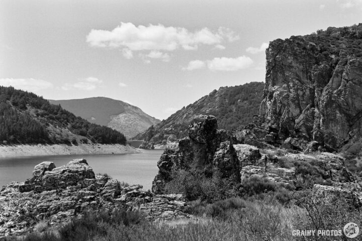 A serene landscape featuring the Camporredondo reservoir surrounded by rocky formations and lush greenery, under a partly cloudy sky. Majestic cliffs rise in the background, showcasing the natural beauty of the area. The image is in black and white, enhancing its artistic feel.