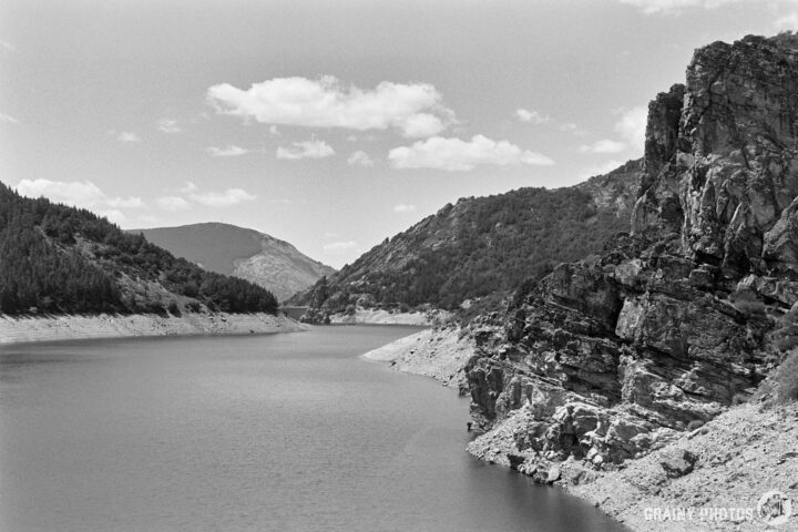 A serene black and white landscape featuring a calm reservoir bordered by rocky cliffs and lush mountains under a partly cloudy sky. The peaceful scene captures the beauty of nature's harmony.