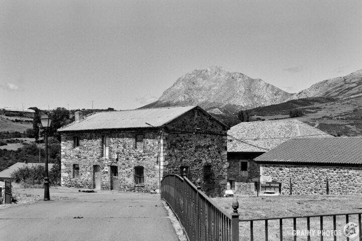 A scenic black and white view of a rustic stone house beside a road, with a mountainous backdrop and lush greenery surrounding the area. A lamp post stands nearby, adding charm to the countryside landscape.