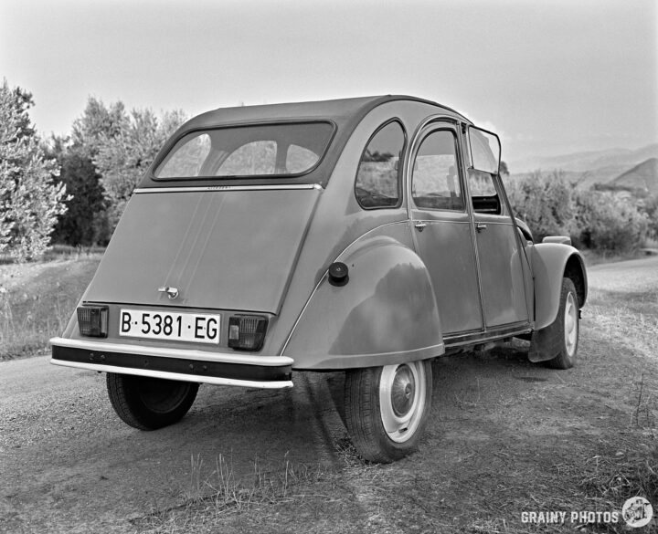 A classic Citroen 2CV car parked on a gravel road, showcasing its rear profile. The vehicle has a rounded shape and a distinct design typical of classic models, surrounded by greenery and distant hills. The image is in black and white, giving it a nostalgic feel.