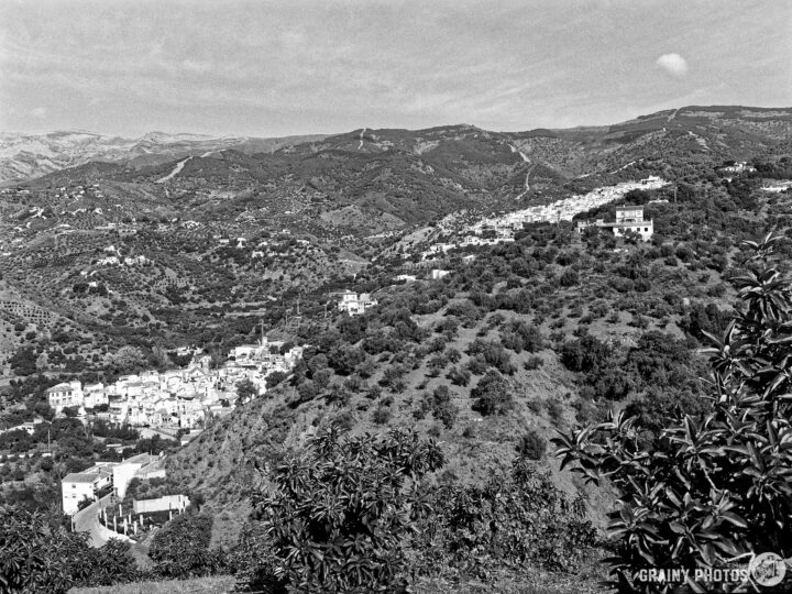A panoramic black-and-white view of a mountainous landscape featuring the villages of Árchez and Canillas de Albaida nestled within lush greenery, surrounded by rolling hills and distant peaks under a clear sky.