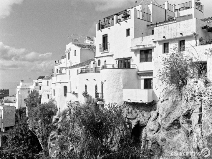 A scenic view of whitewashed buildings perched on a rocky cliff, surrounded by greenery, under a cloudy sky. The composition showcases the blend of nature and architecture in a picturesque setting.