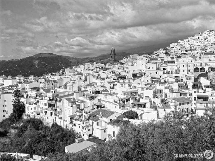 A panoramic view of a Cómpeta on a hillside, with terraced houses and a church tower rising in the background, under a cloudy sky. The scene captures the essence of Mediterranean architecture and landscape in black and white.