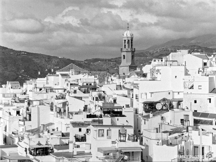 A panoramic view of a Cómpeta featuring the bell tower of Parroquia Nuestra Señora de la Asunción in the background, surrounded by hilly terrain and cloudy skies, showcasing the architectural beauty and charm of Mediterranean-style buildings.