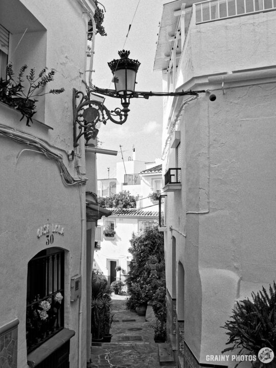 A narrow alleyway lined with white buildings and greenery, featuring a decorative lantern. The scene captures the charm of a quaint village with cobblestone paths and an inviting atmosphere, creating a serene and picturesque setting.