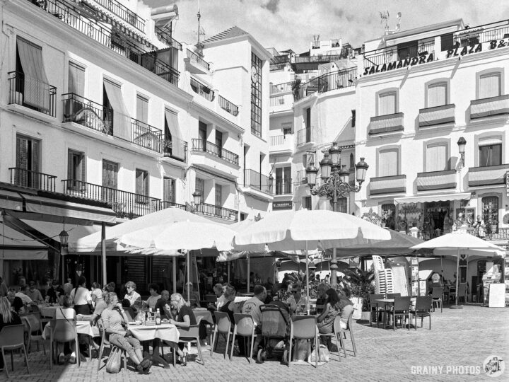 A bustling square filled with people dining under large umbrellas, surrounded by white buildings with balconies. The scene captures a lively atmosphere in a popular outdoor café setting, highlighting social interactions and leisure in a charming environment.