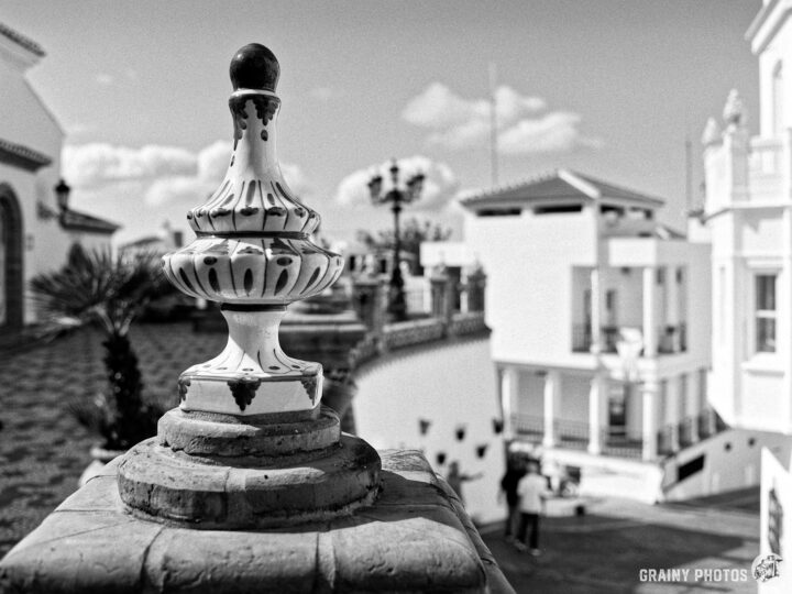A decorative ceramic finial in the foreground by Paseo de las Tradiciones in Cómpeta, with a blurred background featuring white buildings, palm trees, and a clear sky, creating a serene architectural scene in black and white.