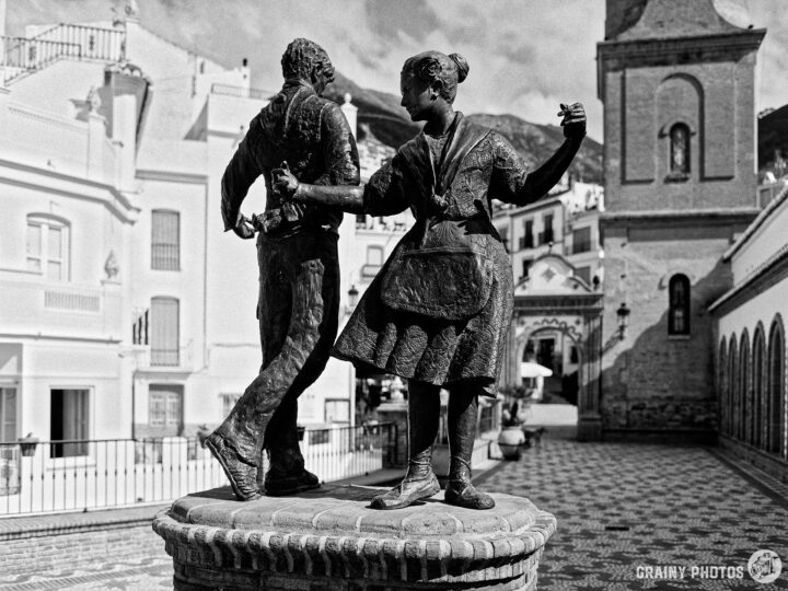 A black and white photo of a bronze sculpture depicting a man and woman dancing joyfully in the picturesque Paseo de las Tradiciones, surrounded by traditional architecture and cobblestone paths, capturing a lively cultural moment.