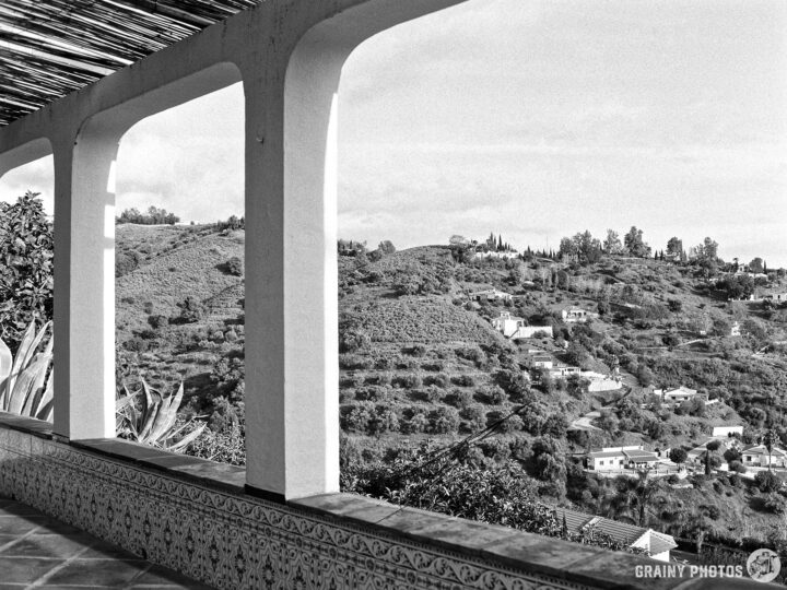 Black and white image of a scenic valley view near Cómpeta from a covered terrace, featuring rolling hills, scattered houses, and lush greenery framed by white pillars and decorative tiles.