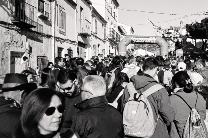 A crowded street scene in black and white, featuring a large group of people ready for the start of the Cross Pantano del Víboras, with buildings in the background and a festive archway across the start-line. The mood is lively, capturing the energy of a community gathering.