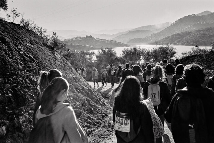 A group of hikers walking along a dirt path, surrounded by hills and trees, with a lake in the background. The scene is captured in black and white, emphasizing the nature of the hike and the scenic landscape.